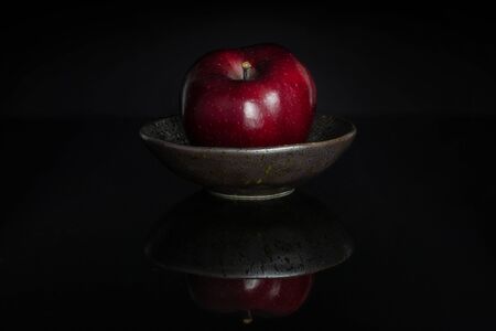 One whole fresh apple red delicious in a dark ceramic bowl isolated on black glassの写真素材