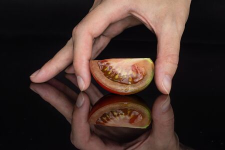One slice of fresh greenish red tomato with hand isolated on black glassの写真素材