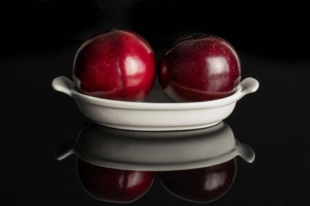Group of two whole ripe red round plum in white oval ceramic bowl isolated on black glassの写真素材