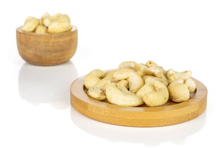 Lot of whole unsalted beige cashew on round bamboo coaster in tiny wooden bowl isolated on whiteの写真素材