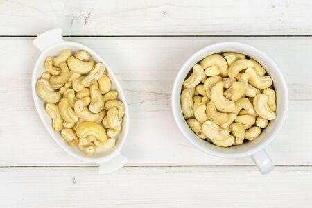 Lot of whole unsalted beige cashew in white oval ceramic bowl flatlay on white woodの写真素材