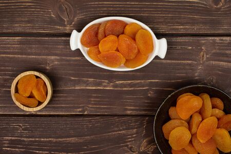 Lot of whole dried orange apricot in tiny wooden bowl in white oval ceramic bowl in dark ceramic bowl flatlay on brown woodの写真素材