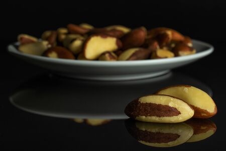 Lot of whole raw brown brazil nut on white ceramic plate isolated on black glassの写真素材