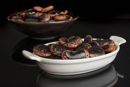 Lot of whole stained purple bean in white oval ceramic bowl in dark ceramic bowl isolated on black...