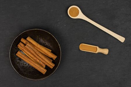 Lot of whole dry brown cinnamon in dark ceramic bowl in a wooden scoop in a wooden spoon flatlay on grey stoneの写真素材