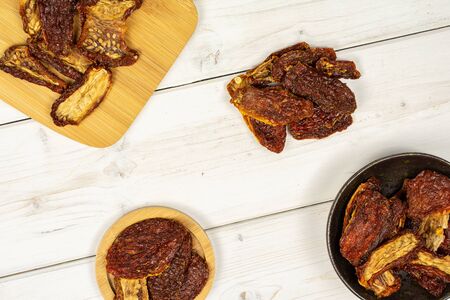 Lot of whole dried red tomato in dark ceramic bowl on round bamboo coaster on bamboo cutting board flatlay on white woodの写真素材