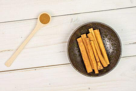 Lot of whole dry brown cinnamon in dark ceramic bowl in a wooden spoon flatlay on white woodの写真素材