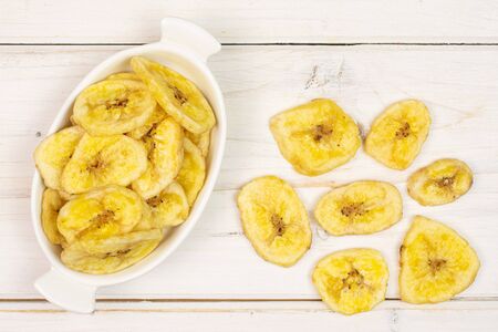 Lot of slices of sweet yellow dry banana in white oval ceramic bowl flatlay on white woodの写真素材
