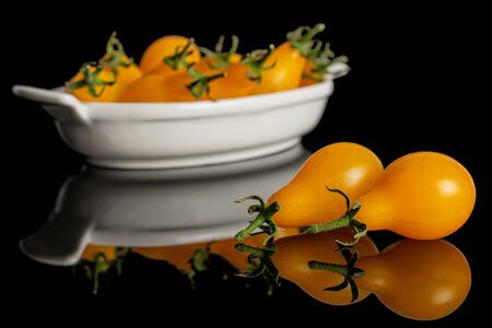 Lot of whole fresh yellow pear tomato two are aside and the rest is in white oval ceramic bowl isolated on black glassの写真素材