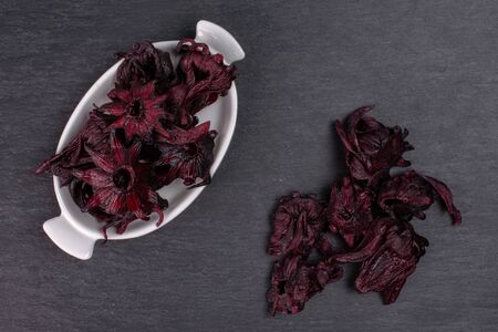Lot of whole sweet red candied hibiscus in white oval ceramic bowl flatlay on grey stone