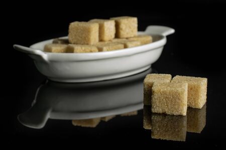 Lot of whole sweet brown sugar cube in white oval ceramic bowl isolated on black glassの写真素材
