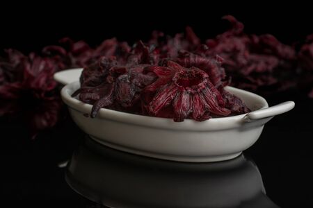 Lot of whole sweet red candied hibiscus in white oval ceramic bowl isolated on black glassの写真素材