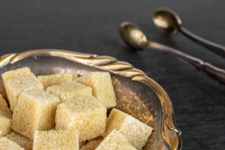 Lot of whole sweet brown sugar cube in metal bowl with sugar tongs on grey stoneの写真素材