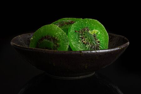 Group of three slices of sweet green candied kiwifruit in dark ceramic bowl isolated on black glassの写真素材