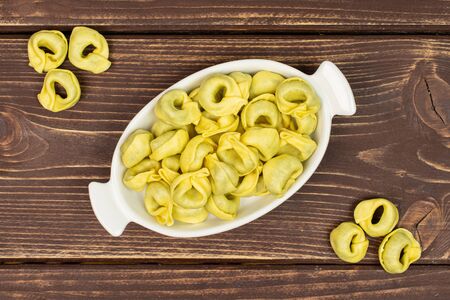 Lot of whole fresh yellow spinach filled tortelloni in white oval ceramic bowl flatlay on brown woodの写真素材