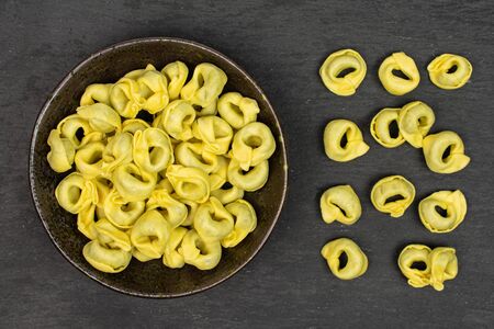 Lot of whole fresh yellow spinach filled tortelloni in dark ceramic bowl flatlay on grey stoneの写真素材