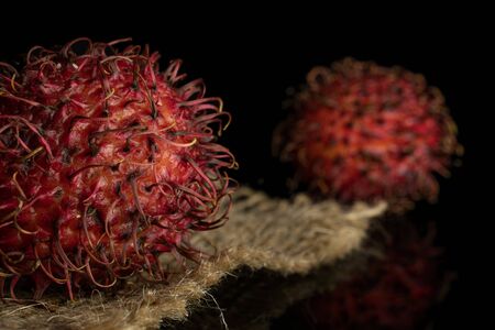 Group of two whole fresh red rambutan closeup on jute cloth isolated on black glassの写真素材