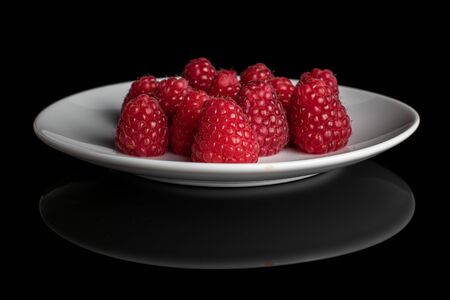 Lot of whole fresh red raspberry on white ceramic plate isolated on black glass
