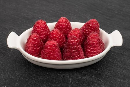 Group of nine whole fresh red raspberry in white oval ceramic bowl on grey stoneの写真素材