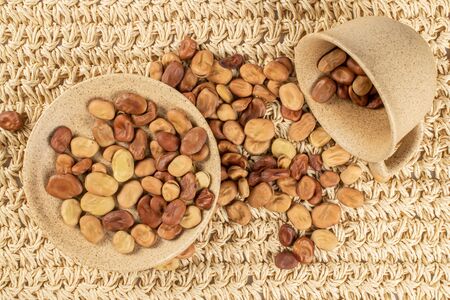 Lot of whole fresh brown bean vicia faba with ceramic saucer and ceramic cup flatlay on straw mat