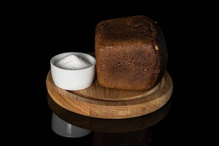 One whole fresh baked dark bread on bamboo plate with pinch of salt isolated on black glassの写真素材