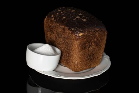 One whole fresh baked dark bread on white plate with pinch of salt isolated on black glass