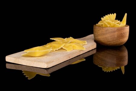 Lot of whole yellow pasta farfalle in bamboo bowl on wooden cutting board isolated on black glass