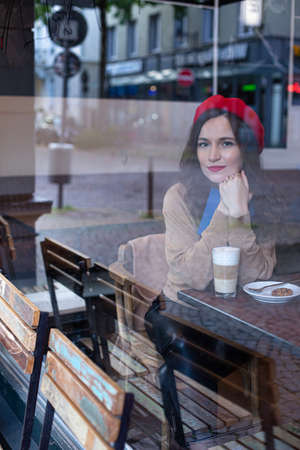 Pretty woman with curly hair in red french beret outside the cafe window drinking latte and holding...