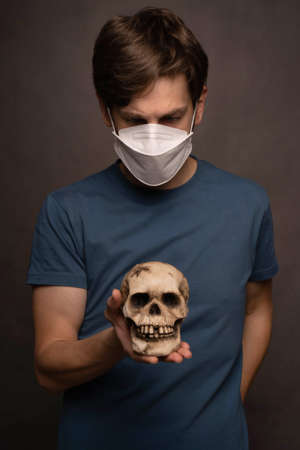 Young handsome tall slim white man with brown hair looking down at skull wearing face mask in blue shirt on grey backgroundの写真素材
