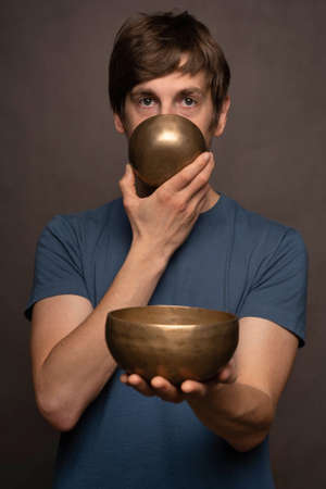 Young handsome tall slim white man with brown hair holding bowl and covering mouth with another bowl in blue shirt on grey backgroundの写真素材