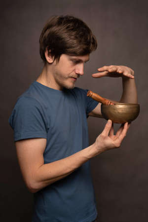 Young handsome tall slim white man with brown hair focusing on tibetan singing bowl in blue shirt on grey backgroundの写真素材