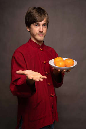 Young handsome tall slim white man with brown hair reaching with his hand holding oranges in red shirt on grey backgroundの写真素材