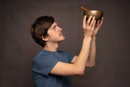 Young handsome tall slim white man with brown hair worshipping tibetan singing bowl in blue shirt on...