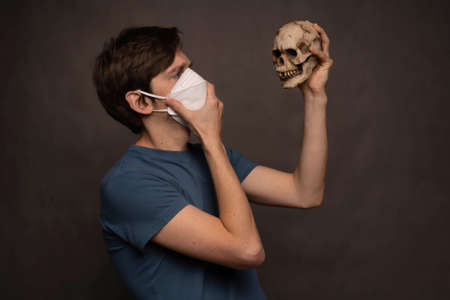 Young handsome tall slim white man with brown hair thoughtful holding skull wearing face mask in blue shirt on grey backgroundの写真素材
