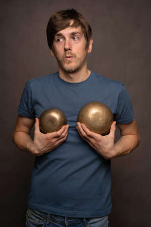 Young handsome tall slim white man with brown hair using bowls as bra in blue shirt on grey...