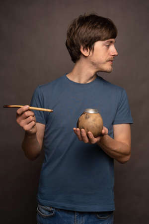 Young handsome tall slim white man with brown hair holding mate cup facing right in blue shirt on grey backgroundの写真素材