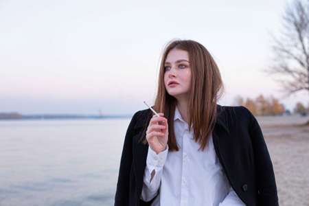 Smoking long straight haired white european woman in dark coat in autumn on the beach