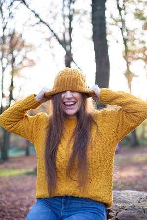 Laughing white young european woman in yellow sweater in sunny autumn forest pulled hat over her eyesの写真素材