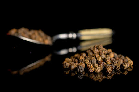 Lot of whole spicy black pepper with metal spoon isolated on black glass