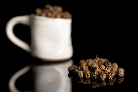 Lot of whole spicy black pepper in small ceramic cup isolated on black glassの写真素材