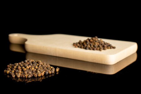 Lot of whole spicy black pepper on small cutting board isolated on black glassの写真素材