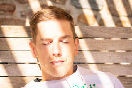 Young handsome white man with brown hair in white shirt is resting outside with closed eyes with sun...