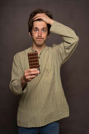 Young handsome tall slim white man with brown hair holding chocolate bar looking worried in striped...