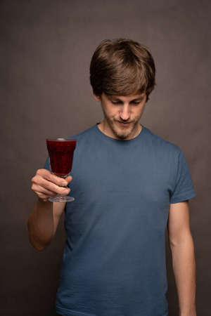 Young handsome tall slim white man with brown hair holding wine cup facing down in grey shirt on...