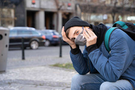 Young handsome white man with brown hair in blue jacket with black winter hat looks desperate holding his head on the streetの写真素材