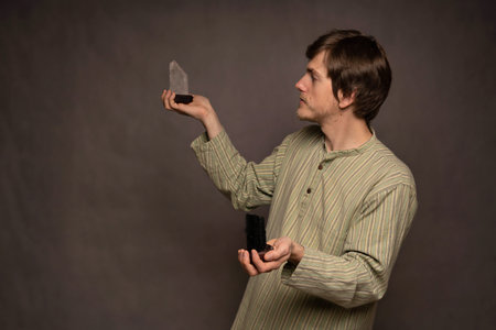 Young handsome tall slim white man with brown hair holding black and white crystals observing the white one in striped shirt on grey backgroundの写真素材