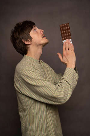 Young handsome tall slim white man with brown hair praying with chocolate bar in his hands in striped shirt on grey backgroundの写真素材