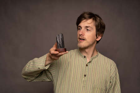 Young handsome tall slim white man with brown hair observing black crystal in striped shirt on grey...