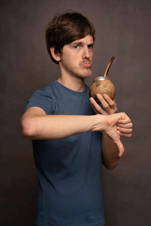 Young handsome tall slim white man with brown hair holding mate cup giving thumbs down in grey shirt on grey backgroundの写真素材