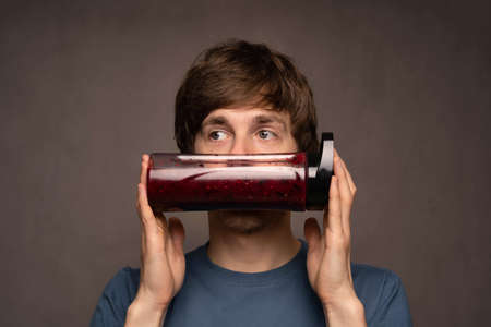 Young handsome tall slim white man with brown hair covering mouth with smoothie bottle in grey shirt on grey backgroundの写真素材